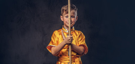 Cute boy dressed in the traditional costume of a Tibetan monk posing with dao stick at the studio.