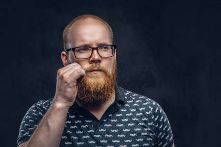 Portrait of a redhead bearded male dressed in a t-shirt posing with a hand on his mustache. Isolated on a dark textured background.の写真素材