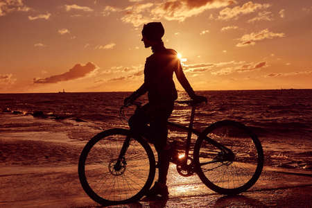 Silhouette of a girl with a bicycle on the sea coast on a sunset background.の写真素材