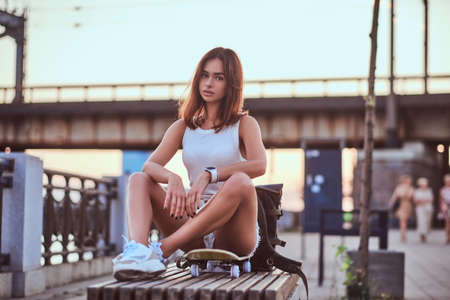 Young sensual skater girl dressed in shorts and t-shirt sitting on the embankment during beautiful sunset.の写真素材