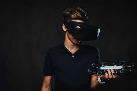 Young teenager dressed in a black t-shirt wears virtual reality glasses and holds a quadcopter....
