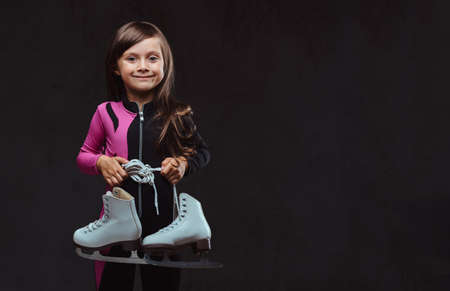 Smiling little girl dressed in sportswear holds ice skates. Isolated on a dark textured background.の写真素材