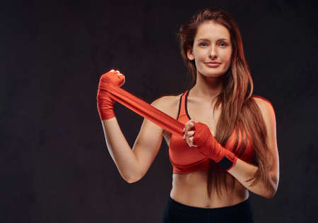 Smiling beautiful brunette female boxer in sports bra bandages her hands. Isolated on a dark...