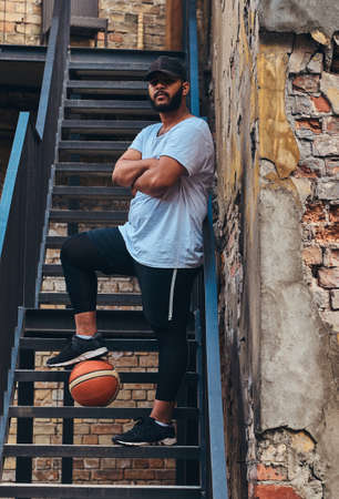 African-American bearded streetball player in cap dressed in a sportswear holds a basketball standing with crossed arms on stairs in ghetto.の写真素材