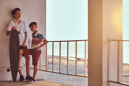 Two little brothers standing with a skateboard near guardrail against the background of the seacoast at a sunset.の写真素材