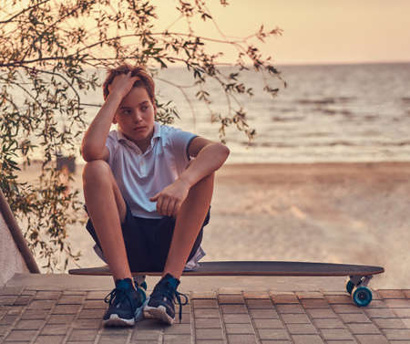 Young skater boy sitting on a skateboard against the background of a seacoast at a sunset.の写真素材