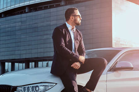 Handsome bearded male in sunglasses dressed in a black suit sitting on luxury car against a skyscraper.の写真素材