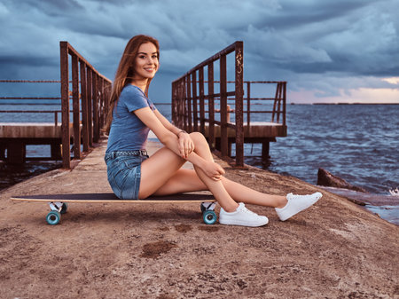 Sensual girl sitting on a skateboard on the beach is enjoying amazing dark cloudy weather during...