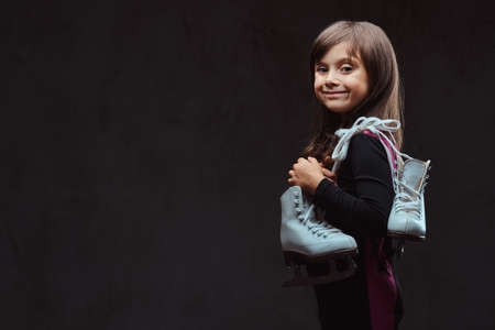 Smiling little girl dressed in sportswear holds ice skates on a shoulder. Isolated on dark textured background.の写真素材