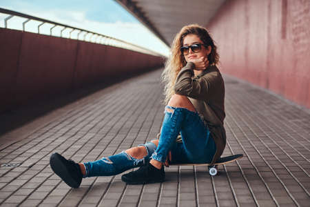 Portrait of a pensive girl in sunglasses dressed in a hoodie and ripped jeans sitting on a skateboard at bridge footway.の写真素材