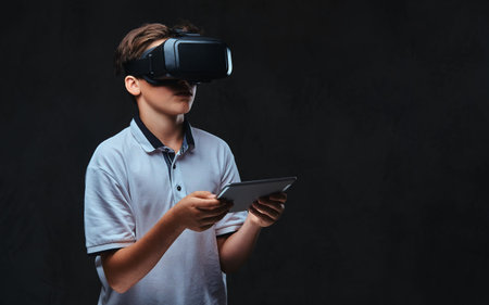 Portrait of a young student boy dressed in a white t-shirt using virtual reality glasses and tablet computer.の写真素材