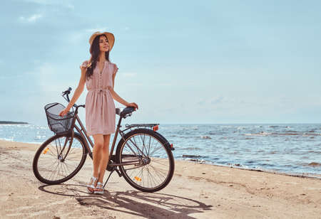 Beautiful brunette girl dressed in dress and hat posing with a bicycle on the beach on a sunny day.の写真素材