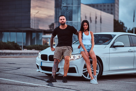 Cool couple - brutal tattooed male and sensual girl leaning on a luxury car outdoors against the...