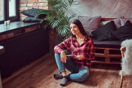 Charming brunette girl student dressed in a plaid shirt and jeans holding digital tablet sitting on a floor in a room with loft interior.の写真素材