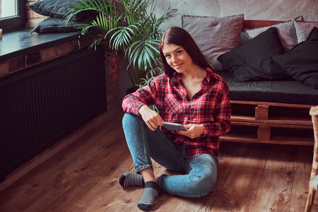 Charming brunette girl student dressed in a plaid shirt and jeans holding digital tablet sitting on a floor in a room with loft interior.の写真素材