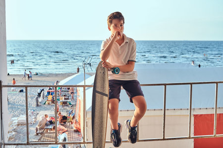 Portrait of a young skater boy dressed in t-shirt and shorts sitting on the guardrail and holds a skateboard against the background of a seacoast.の写真素材