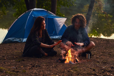 Happy young couple sitting and warming near a campfire at camp in the forest at sunset.の写真素材