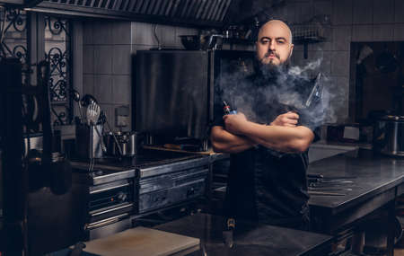 Bearded chef in black uniform smoking e-cigarette while standing in the kitchen.の写真素材