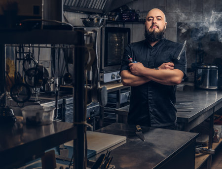 Bearded chef in black uniform smoking e-cigarette while standing in the kitchen.