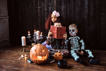 Group of cute multiracial kids in scary costumes reading horror stories in an old house, during Halloween party.の写真素材