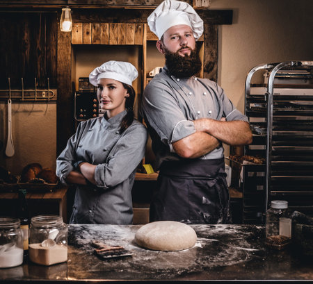 Chef with his assistant in cook uniform posing with crossed arms near table with ready dough in the bakery.の写真素材