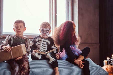 Three multiracial kids in scary costumes reading horror stories while sitting on bed in an old...