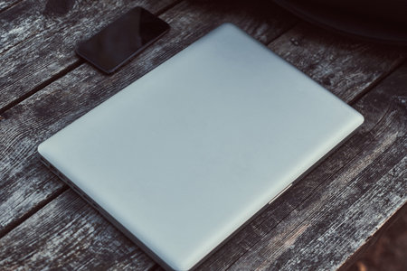Close-up photo of a laptop and smartphone on wooden table outdoors.