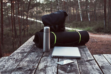 Travel concept. Backpack, tourist mat, laptop, smartphone and thermos bottle on a wooden table in beautiful forest.の写真素材