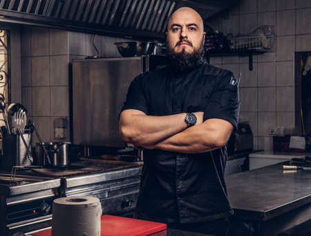 Brutal bearded chef cook in black uniform standing with crossed arms in the kitchen.の写真素材