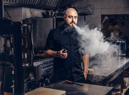 Bearded chef in black uniform smoking e-cigarette while standing in the kitchen.の写真素材