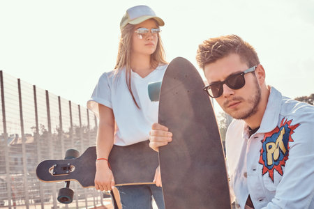 Close-up portrait of young trendy dressed hipster couple with skateboards posing at city sports...