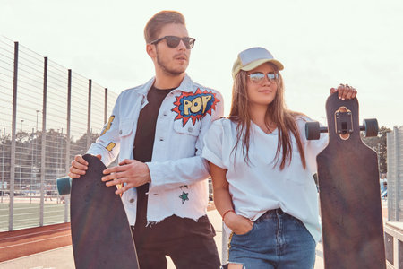 Atractive couple of trendy dressed young hipsters posing with skateboards at city sports complex on...