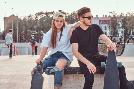 Young stylish couple of a trendy dressed skaters relaxing at a skatepark.