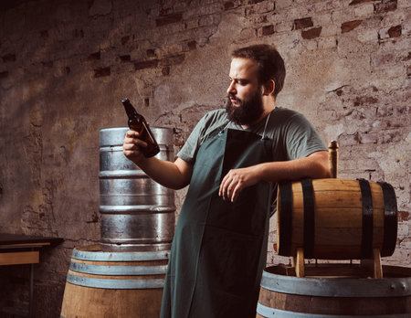 Brewer in apron standing near barrels and drinks craft beer at old brewery factory.の写真素材