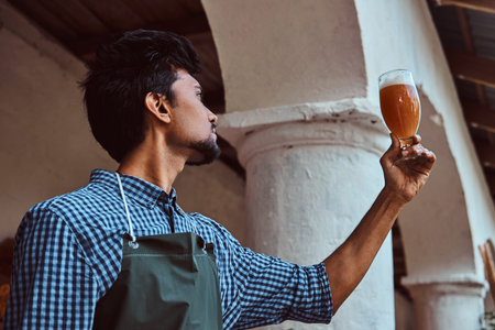 Expert brewer in apron standing outdoors checking the quality of brewed drink at brewery factory.