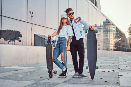 Attractive young couple dressed in trendy clothes posing with skateboards near skyscraper.