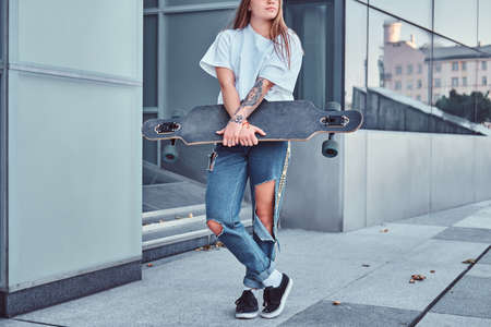 Cropped photo of a young hipster girl dressed in white shirt and ripped jeans holds skateboard while posing near skyscraper.の写真素材