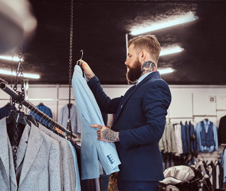Elegantly dressed bearded male with tattoos on hands and neck chooses new costume in the menswear...