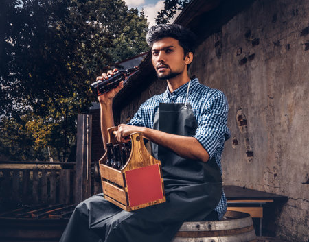 Brewmaster sitting on a wooden barrel and holds a glass of craft beer, relaxes after work.