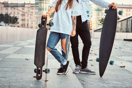Cropped photo of a trendy dressed young couple posing with skateboards near a skyscraper.の写真素材