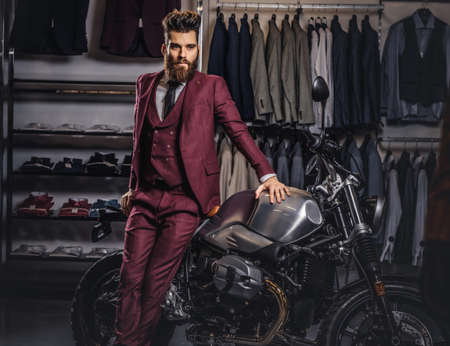 Handsome man with a stylish beard and hair dressed in vintage red suit posing near retro sports motorbike at mens clothing store.の写真素材