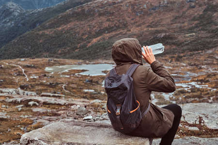 Back view of a tourist who drinks water and sitting on a rock in the background of a beautiful mountain landscape.の写真素材