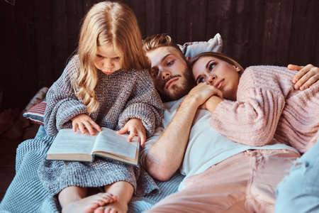 Mom, dad and daughter reading storybook together while lying on bed.