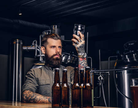 Male with stylish beard and hair holds pint of craft beer sitting at the bar counter in the indie brewery.の写真素材