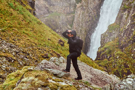 Nature photographer tourist with camera shoots while standing on the mountain against a waterfall.の写真素材