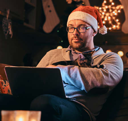 Man in Santa hat and glasses dressed in warm sweater holds cat and using laptop celebrating Christmas at home.の写真素材