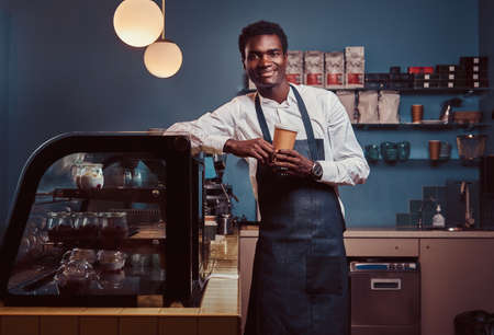 African barista smiling at camera relaxing after workday with coffee while leaning on the counter at coffee shop.の写真素材