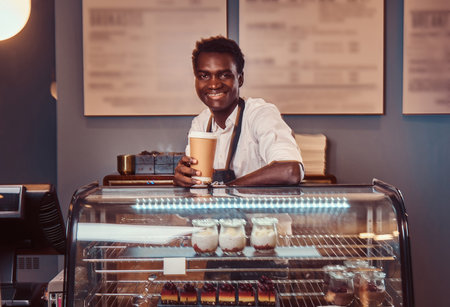 Portrait of a handsome African barista in white shirt and apron holds a cup of coffee and relaxing after workday at coffee shop.の写真素材
