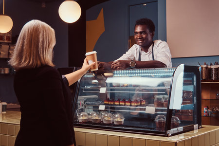 Smiling African American barista in uniform giving a cup of coffee to his client at the trendy...