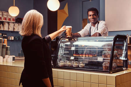 African American barista giving cup of coffee to his female client at the trendy coffee shop.の写真素材
